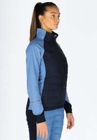 Lightweight, two-tone jacket featuring a dark blue quilted front and light blue stretch sleeves, with a high collar and zip closure.