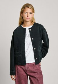 Schwarze Jeansjacke mit rundem Halsausschnitt, fünf silbernen Knöpfen, zwei frontalen Taschen und langen Ärmeln, getragen über einem weißen T-Shirt mit Grafik.