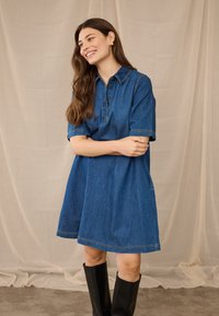 Mosolygó nő hosszú barna hajjal, kék farmerruhát és fekete térdcsizmát visel, bézs színű textil hátteret állva.