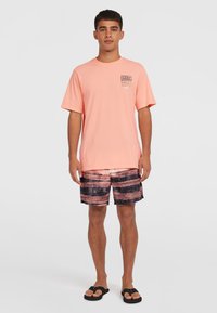 Lyserød t-shirt med korte ærmer og et logo på brystet, parret med mønstrede sorte og ferskenfarvede badebukser. Sorte klipklappere fuldender looket.