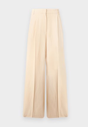 Femme portant un gilet noir boutonné sur une chemise blanche à manches courtes, un pantalon large crème et des sandales noires à bouts ouverts, debout devant un fond blanc.