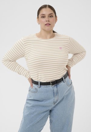 Woman with hands on hips wearing beige striped long-sleeve shirt with small pink heart and light blue high-waisted jeans with black belt.