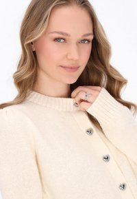 Krémový pletený cardigan s žebrovaným límcem, stříbrnými knoflíčky ve tvaru srdce a měkkou texturou. Modelka drží prsten na prstu.