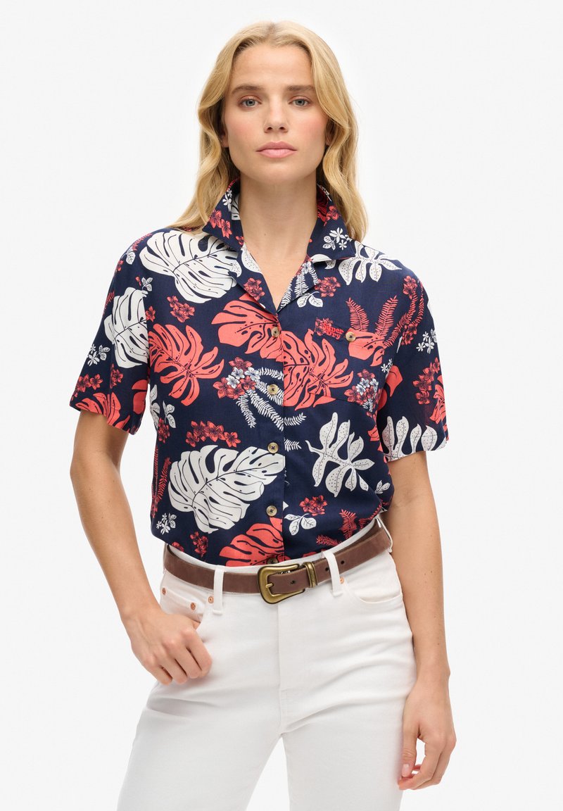 Camisa de manga corta de color azul marino con patrones de hojas y flores tropicales en rojo y blanco. Presenta un cuello y botones marrones, combinada con pantalones blancos.