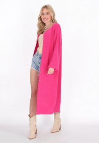 Dolgi roza kardigan z rebrasto teksturo, z zlatimi gumbi, širokimi rokavi in stranskimi razporki, nosi se čez bež majico in jeans kratke hlače.