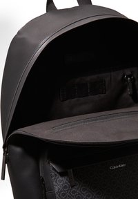 Zaino nero con un'esterno liscio, chiusura con cerniera e una tasca decorata con dettagli a logo. L'interno include una sezione con cerniera.