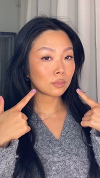 Młoda kobieta z długimi czarnymi włosami, ubrana w szary, puszysty sweter, wskazuje na swoją twarz, na której widać błyszczące usta i subtelny makijaż.