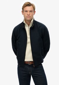 Chaqueta bomber de color azul oscuro con cremallera frontal, cuello redondo y detalles en marrón. Llevada sobre un suéter de cremallera color crema y combinada con jeans azules.
