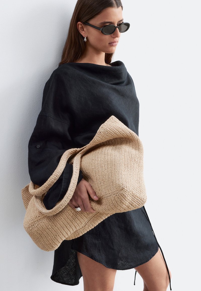 Femme portant une robe noire et des lunettes de soleil, tenant un grand sac cabas beige tricoté contre un mur blanc.