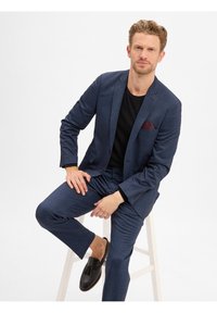 Marineblauwe gestructureerde pak met een slim fit, gecombineerd met een zwart shirt en zwarte instappers; voorzien van een bordeauxrode zakdoek.