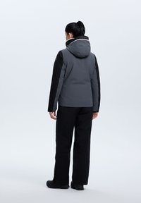 Giacca impermeabile grigia e nera con cappuccio, caratterizzata da una chiusura con zip e polsini elasticizzati. Abbinata a pantaloni neri e calzature robuste.