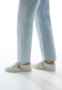 Sneaker bianche in rete con dettagli marrone chiaro, punta rotonda e suola in gomma testurizzata. Abbinati a jeans azzurri dritti.
