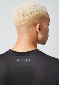 Czarna sportowa koszulka z teksturowaną powierzchnią, z odblaskowym logo "SNOCKS" z tyłu, noszona przez osobę z krótkimi blond włosami.