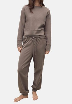 Femme portant un haut marron à manches longues et un pantalon assorti à cordon, debout pieds nus devant un fond clair uni.