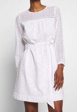 Robe blanche à manches longues, tissu texturé avec des motifs à œillets, taille cintrée avec une ceinture, et jupe droite.
