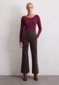 Bordeauxfarbene Langarmbluse kombiniert mit weit geschnittenen, dunkelbraunen Cordhosen und braunen Lederstiefeletten, vor einem neutralen Wandhintergrund.