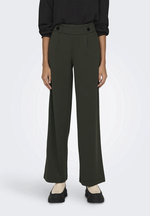 Person, die dunkelgrüne, hoch taillierte, weit geschnittene Hose mit Knopfdetails, schwarzen Langarmtop, cremefarbene Socken und schwarze Plateauschuhe trägt.