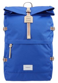 Sac à dos en tissu bleu à rabat roulé avec boucle de sangle beige, sangles latérales réglables, poche avant zippée et étiquette blanche Sandqvist.