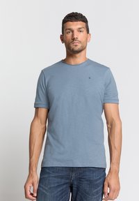 Lichtblauw T-shirt met korte mouwen en ronde hals, zachte textuur, subtiele logo-detail op de borst, gecombineerd met donkerblauwe jeans.