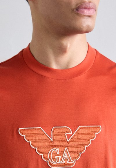 Raudona medvilninė marškinė su apvalia iškirpte, ant krūtinės išsiuvinėtas erelio logotipas oranžinės ir baltos spalvų kombinacijoje.