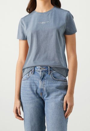 Femme portant un t-shirt bleu délavé Tommy Hilfiger rentré dans un jean en denim bleu clair taille haute, sur un fond uni.