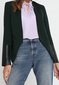 Černý a zelený blazer s dlouhými rukávy a černými umělými koženými manžetami, který je zkombinován s světle fialovou blůzou a džínami s vysokým pasem modré barvy.