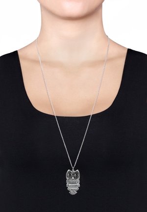 Pendentif chouette en argent avec des yeux en pierre noire suspendu à une longue chaîne portée par une personne vêtue d'un haut noir à encolure dégagée.