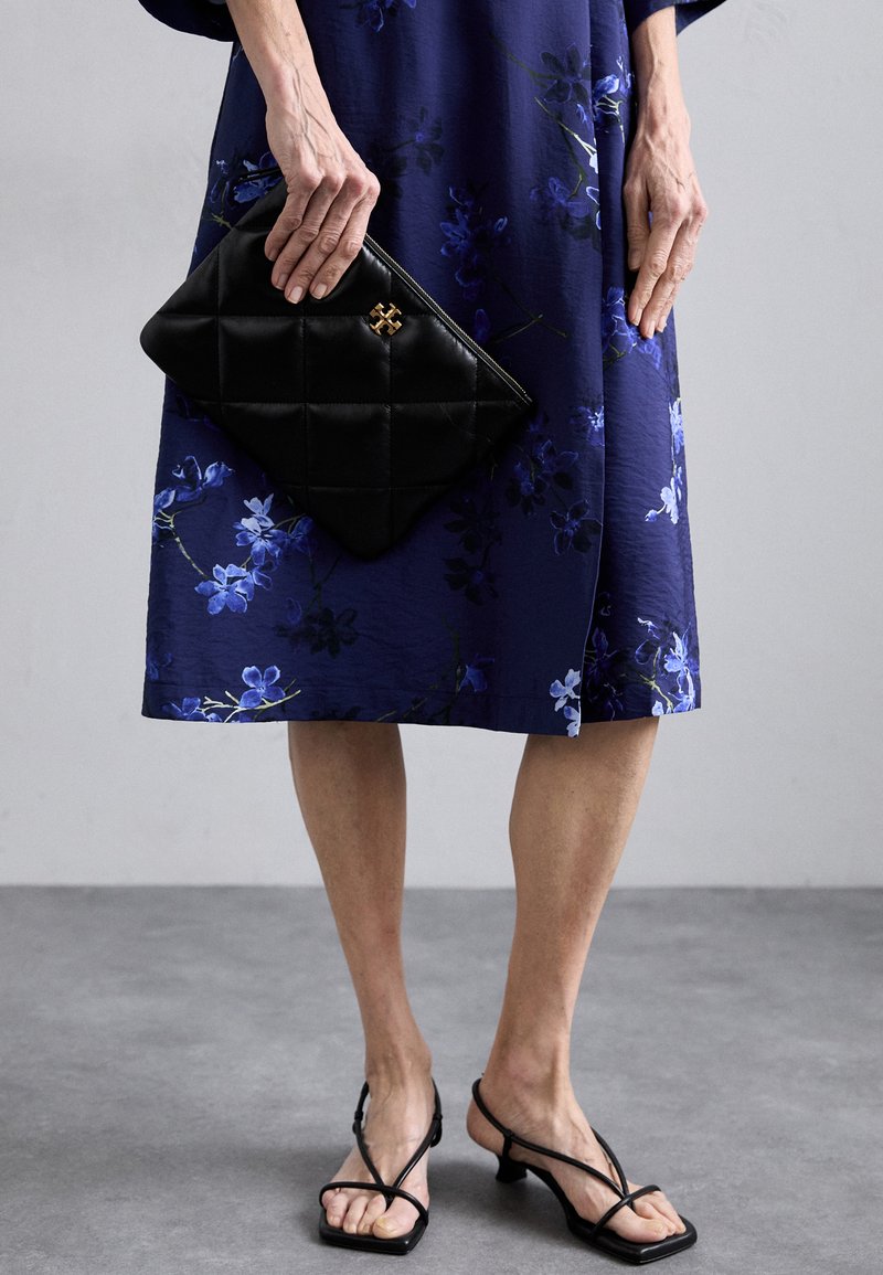 Svart quiltad kuvertväska med guldfärgade detaljer, som kombineras med en blå blommig klänning. Klänningen har en slät textur och knälång silhuett. Svarta remsandaler.