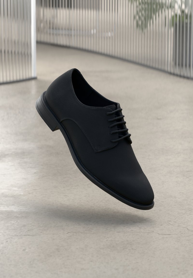 Chaussure habillée pour homme en cuir noir avec lacets, montrée flottant au-dessus d'un sol clair à l'intérieur.