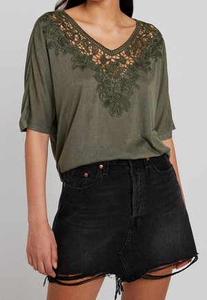 Olijfgroene top met V-hals en gedetailleerd geborduurd bloemenpatroon bij de halslijn, gecombineerd met een zwarte denimrok met rafelige zoom.