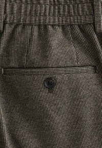 Brązowy materiał w wzór, przedstawiający desen houndstooth. Zawiera przednią kieszeń z czarnym guzikiem. Teksturalny wygląd i poukładany kształt.