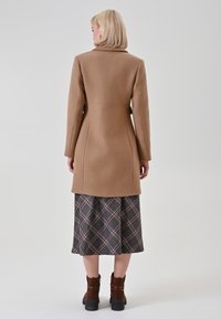 Kamelwollmantel mit einer taillierten Silhouette, ausgestattet mit einem hohen Kragen und langen Ärmeln, kombiniert mit einem karierten Rock und braunen Stiefeletten.