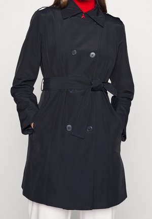 Frau trägt einen schwarzen, doppelreihigen Trenchcoat mit gebundenem Gürtel, die Hände in den Taschen, darunter ein rotes Oberteil.