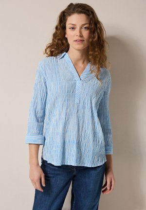 Blouse bleu clair à manches longues avec des rayures blanches verticales, un col en V et des poignets retroussés. Texture douce et coupe ample, associée à un jean bleu.