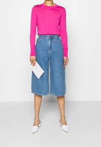 Pull fuchsia à manches longues associé à un pantalon culotte en denim bleu clair. Pochette blanche à la main et mules blanches à bouts pointus aux pieds.