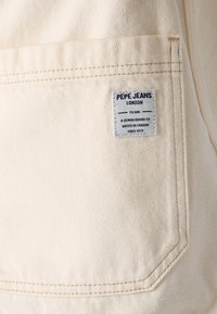 Beiger Denimstoff mit sichtbarer Tasche und einem Logopatch. Das Etikett trägt "Pepe Jeans London" und zeigt Markendetails und ein Nähmuster.