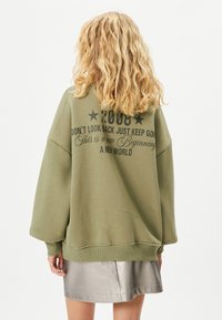 Sudadera de gran tamaño en verde oliva con puños y dobladillo acanalados, que presenta texto impreso en negro y estrellas en la parte posterior. Combinada con una falda plateada.