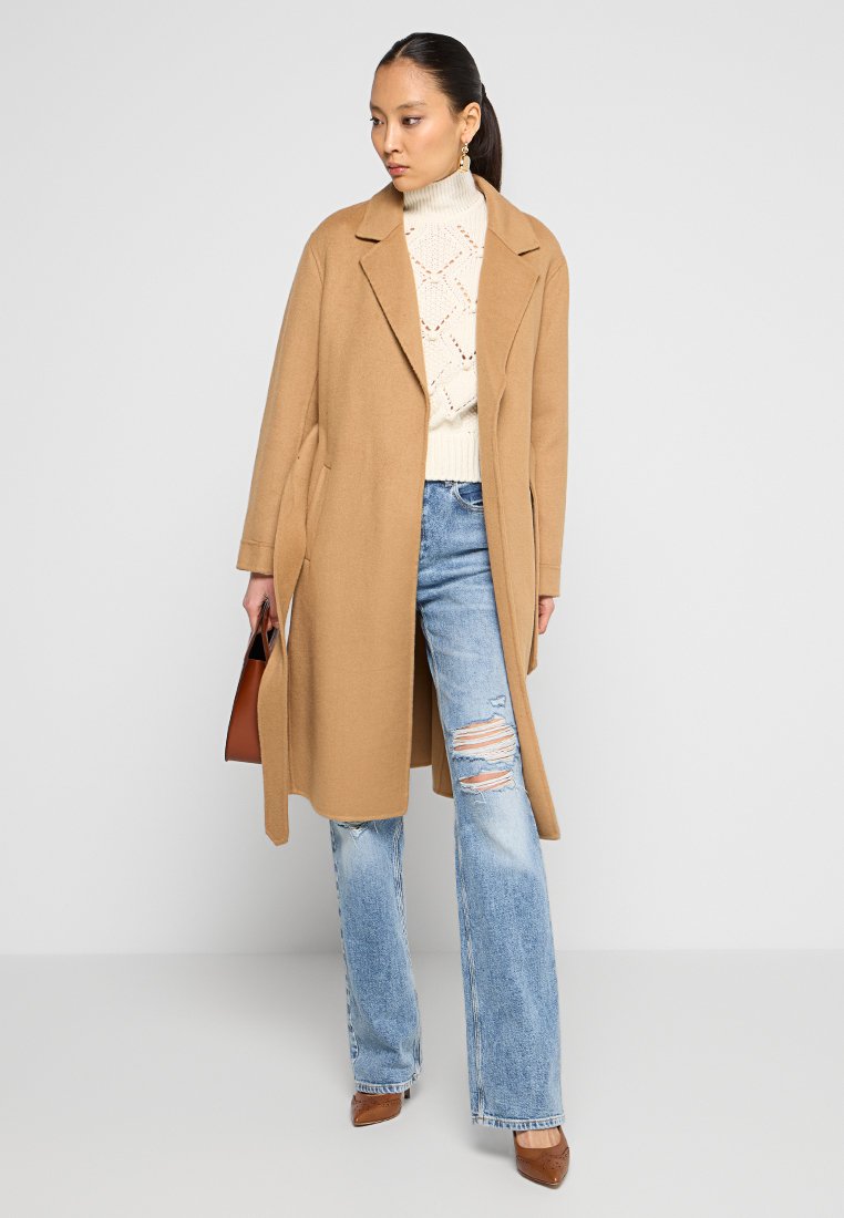 Kasztanowy wełniany płaszcz z wykładanym kołnierzem, długimi rękawami i bocznymi kieszeniami, nałożony na kremowy sweter z dzianiny i przetarte niebieskie dżinsy.