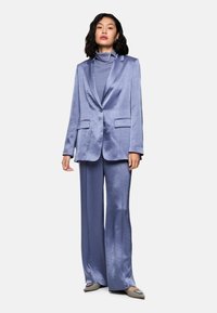 Woman wearing a shiny lavender pantsuit with wide-leg trousers and a high-neck top, standing with neutral expression and gray pointed flats.
