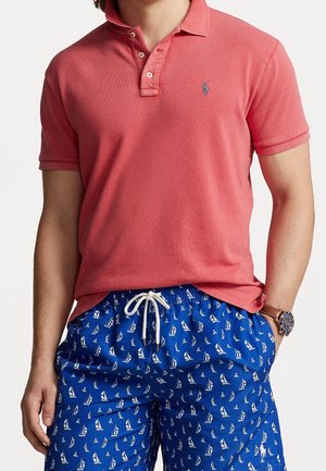 Man in een koraalkleurige polo en blauwe zwembroek met witte zeilbootpatronen, hand in de zak, en een horloge om de linkerpols.