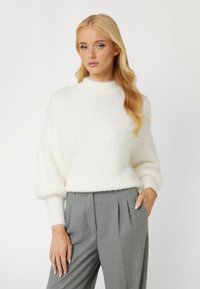 White, textured, fluffy sweater with a high neckline and long sleeves, paired with gray pleated trousers. Simple, clean design.