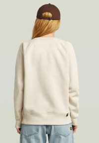 Beige sweatshirt with a crew neck and raglan sleeves, featuring a small tag on the back and cuffed hems. Worn with light blue jeans.