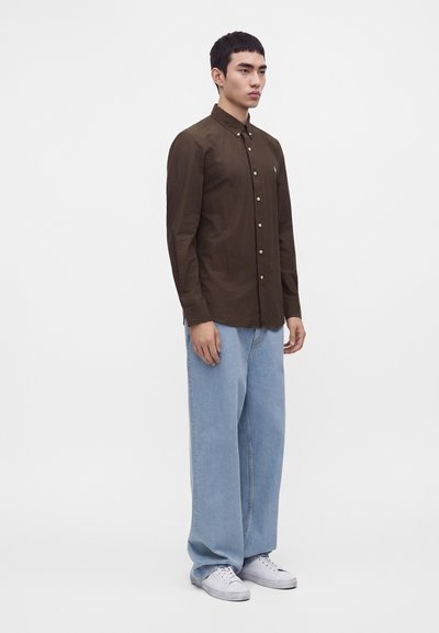 Homme debout portant une chemise boutonnée brun foncé, un jean large bleu clair et des baskets blanches sur un fond blanc uni.
