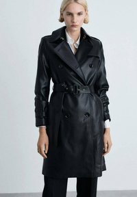 Schwarzer Leder-Trenchcoat mit doppeltreihigen Knöpfen, breiten Revers, tailliertem Schnitt und Bündchendetails, getragen über einem weißen Hemd.