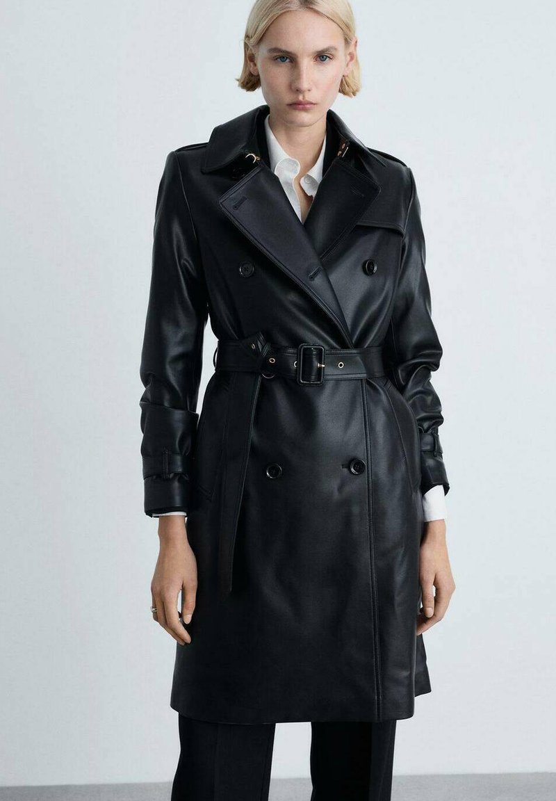 Schwarzer Leder-Trenchcoat mit doppeltreihigen Knöpfen, breiten Revers, tailliertem Schnitt und Bündchendetails, getragen über einem weißen Hemd.