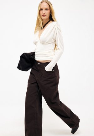 Žena s dlhými blond vlasmi, oblečená v priliehavom bielom nariasenom topi, čiernych širokých nohaviciach, s čiernym korálkovým náhrdelníkom, držiaca čiernu bundu.