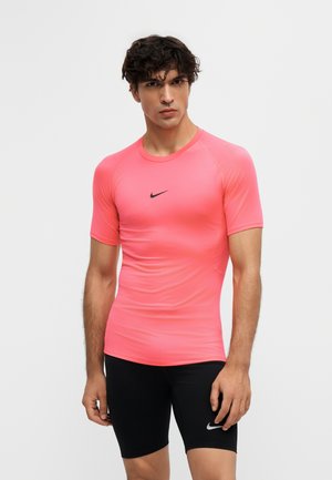 Nuori mies, jolla on tiukka kirkkaan vaaleanpunainen Nike lyhythihainen paita ja mustat Nike urheilushortsit, seisoo yksivärisen valkoisen taustan edessä.