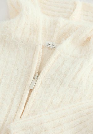 Pull-over crème à fermeture éclair avec une texture douce et moelleuse, un design côtelé et une fermeture éclair argentée. L'étiquette indique "NEXT, Fabriqué au Bangladesh."