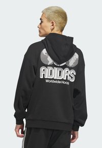 Čierna mikina s kapucňou a veľkým bielym logom "ADIDAS" a textom "Worldwide Hoop" na chrbte, s voľným strihom.