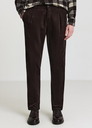 Homme portant un pantalon en velours côtelé brun foncé, des mocassins noirs et une chemise à carreaux noir et blanc, les mains dans les poches.